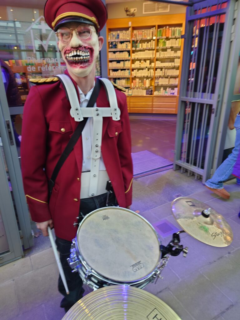 Un batteur déguisé en zombie, portant un masque avec un sourire effrayant, en uniforme rouge, se tient derrière sa caisse claire et ses cymbales.